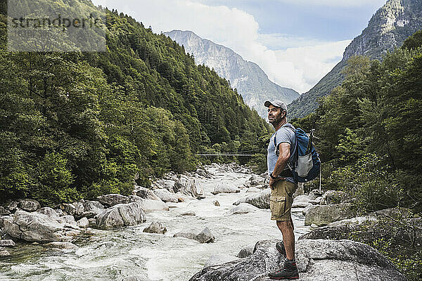 Mature man with hand on hip standing by river in front of mountains