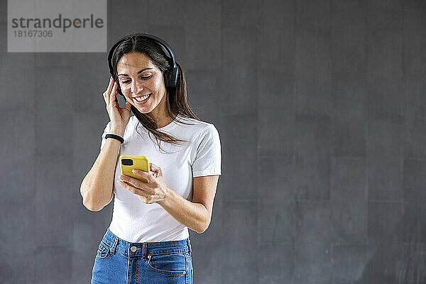 Lächelnde Frau hält Smartphone in der Hand und hört Musik über kabellose Kopfhörer vor grauer Wand