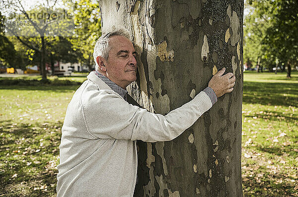 Älterer Mann mit geschlossenen Augen umarmt Baumstamm im Park