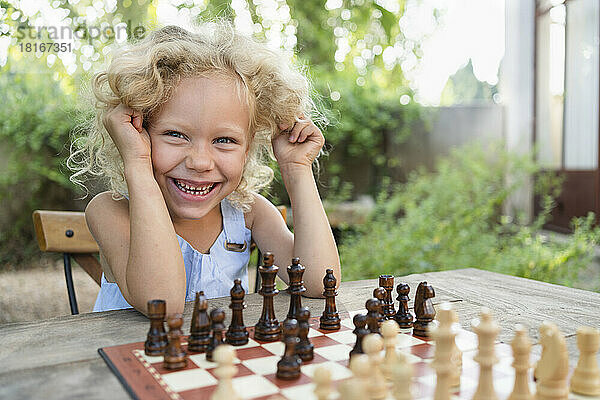 Glückliches blondes Mädchen sitzt mit Schachbrett im Garten