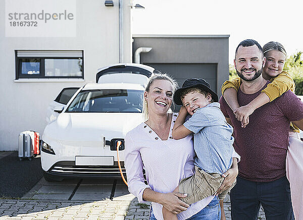 Happy family standing in front of family home and charging electric car