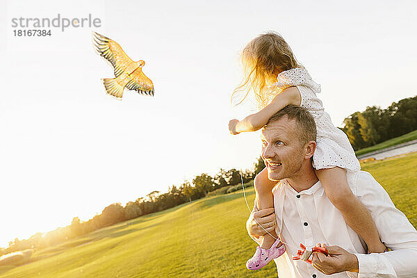 Vater trägt Tochter auf der Schulter und lässt Drachen im Park steigen