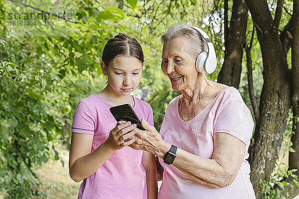 Mädchen hilft Urgroßmutter mit Smartphone am Baum im Park