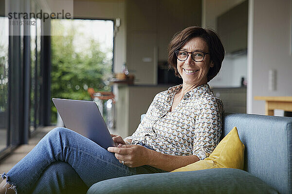 Glückliche Frau mit Laptop sitzt zu Hause auf dem Sofa