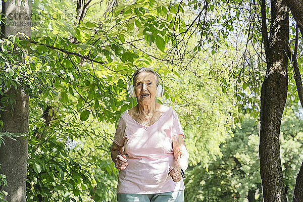 Glückliche ältere Frau mit kabellosen Kopfhörern joggt inmitten von Bäumen