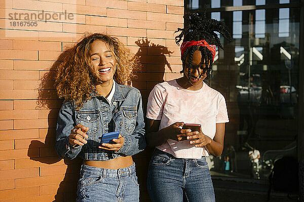 Junge Frau lacht an einem sonnigen Tag mit einer Freundin  die ihr Mobiltelefon benutzt