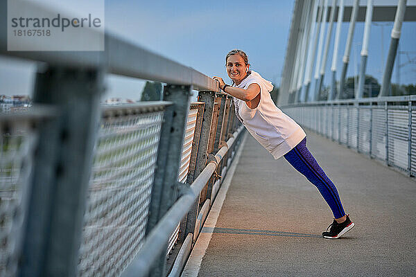 Lächelnde Frau trainiert in der Nähe des Geländers an der Brücke