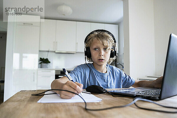 Nachdenklicher Junge mit kabelgebundenen Kopfhörern  der zu Hause am Laptop lernt