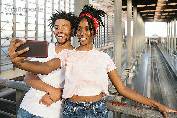 Lächelnder junger Mann und junge Frau  die am Bahnhof ein Selfie mit dem Smartphone machen