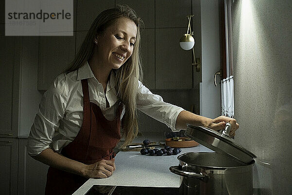 Lächelnde Frau öffnet den Deckel eines Topfes in der Küche