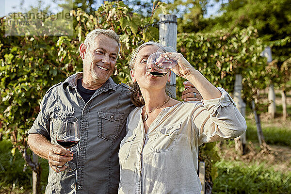 Lächelnder reifer Mann mit Weinglas und Blick auf eine Frau  die Wein vor dem Weinberg trinkt