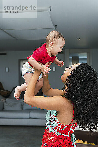 Glückliche Frau mit lockigem Haar hält ihren Sohn zu Hause hoch