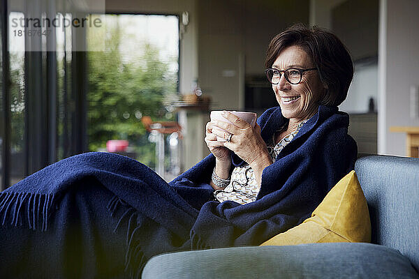 Glückliche ältere Frau  in eine Decke gehüllt  sitzt mit einer Kaffeetasse auf dem Sofa zu Hause