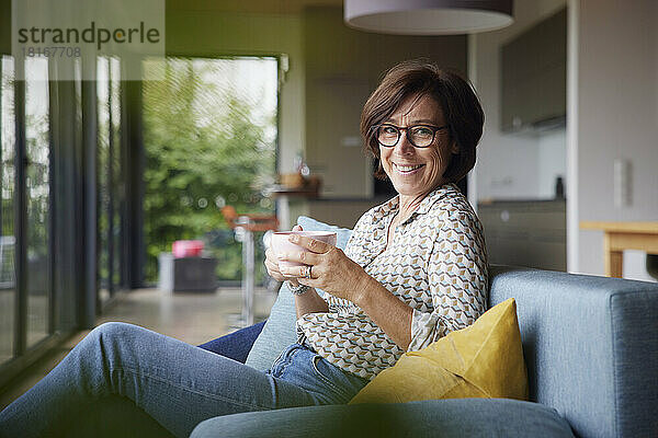 Glückliche Frau mit Kaffeetasse sitzt zu Hause auf dem Sofa