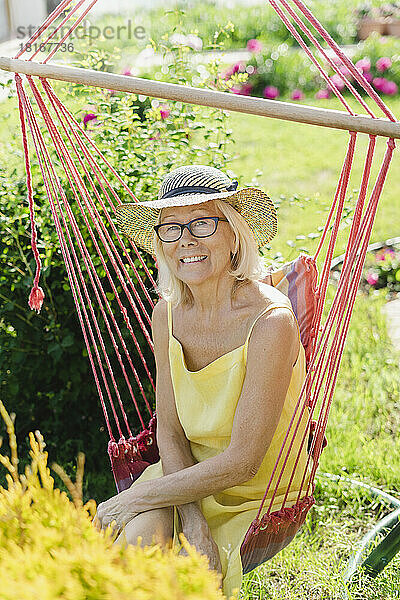 Lächelnde ältere Frau mit Hut sitzt auf einem Hängesessel im Garten