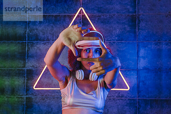 Junge Frau macht Fingerrahmen vor dreieckigem Neonlicht an der Wand