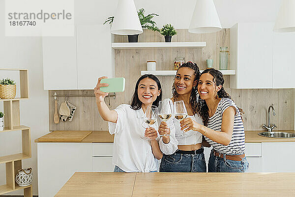 Happy friends talking selfie with wineglasses in kitchen
