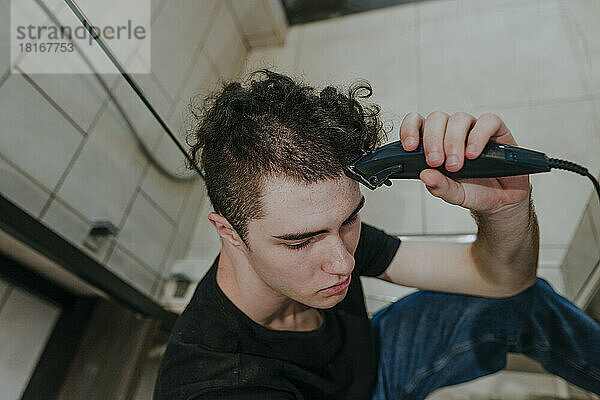 Trauriger junger Mann mit Elektrorasierer  der sich im Badezimmer die Haare rasiert