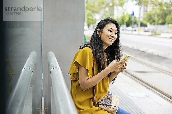 Lächelnde Frau mit Mobiltelefon an der Straßenbahnhaltestelle