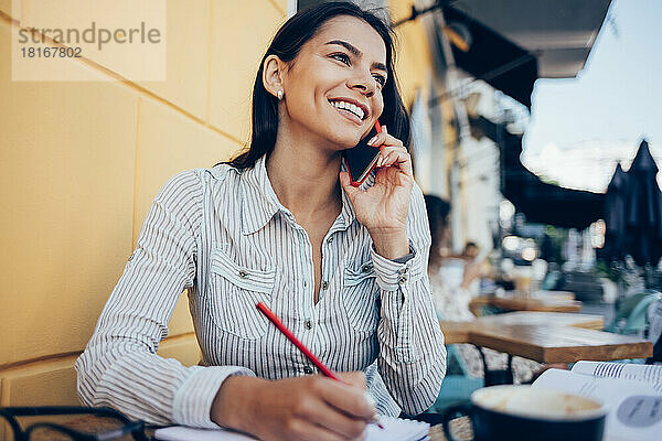 Glückliche Geschäftsfrau  die im Straßencafé mit dem Smartphone spricht