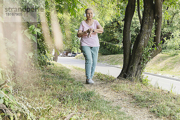 Aktive Seniorin mit kabellosen Kopfhörern geht im Park an Baum vorbei