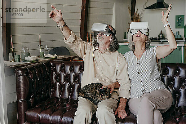 Mann und Frau mit VR-Brille gestikulieren mit Katze auf dem Schoß zu Hause