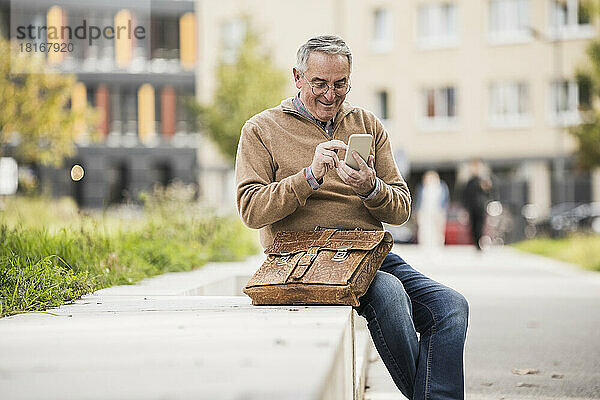 Lächelnder Mann benutzt Smartphone und sitzt mit Tasche auf Betonbank