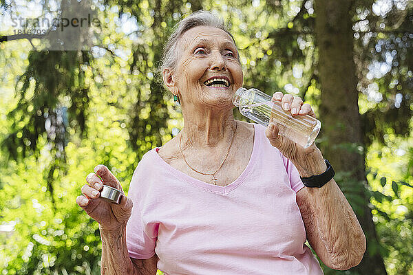 Glückliche ältere Frau mit Wasserflasche  die vor einem Baum im Park steht