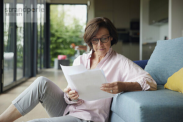 Ältere Frau liest Dokumente und sitzt zu Hause auf dem Sofa