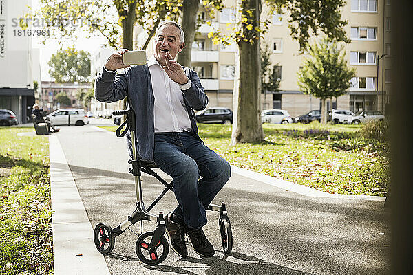 Älterer Mann gestikuliert bei Videoanruf  während er über sein Smartphone sitzt und auf einem Rollator sitzt