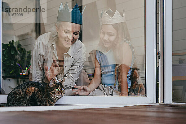 Glückliche Enkelin und Großmutter mit Krone und Blick auf die Katze durch das Glasfenster