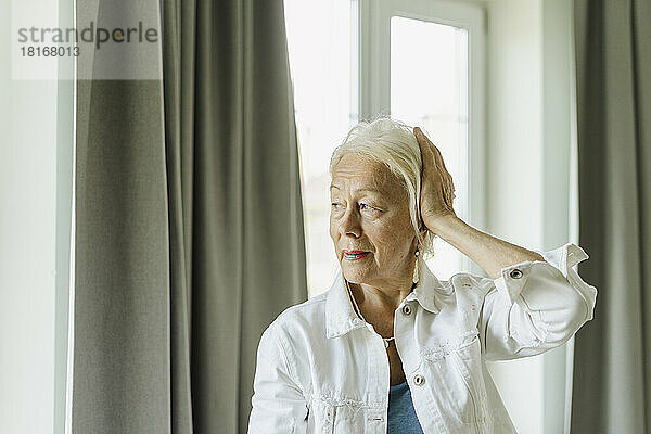 Ältere Frau berührt weißes Haar in der Nähe des Vorhangs zu Hause