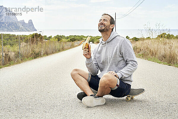 Fröhlicher Mann mit Banane sitzt auf Skateboard