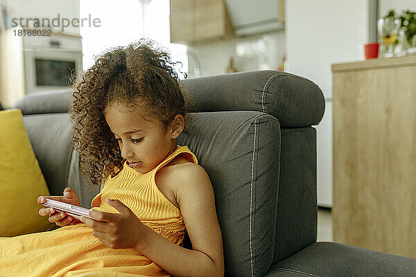 Mädchen mit lockigem Haar schaut sich zu Hause auf dem Sofa ein Video per Handy an