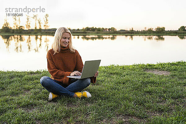 Blonder Freiberufler arbeitet am Laptop und sitzt mit gekreuzten Beinen im Gras am See