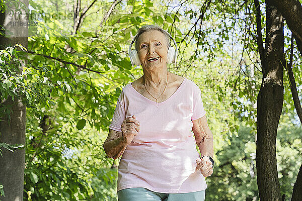 Glückliche aktive Seniorin mit kabellosen Kopfhörern joggt inmitten von Bäumen