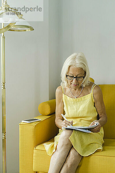 Ältere Frau schreibt in einem Buch und sitzt zu Hause auf dem Sofa