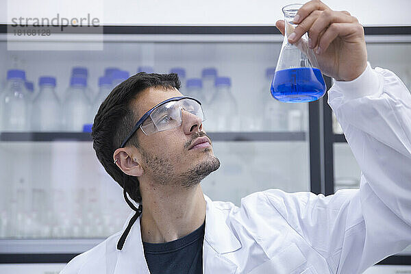 Wissenschaftler mit Schutzbrille untersuchen blaue Chemikalie im Kolben im Labor