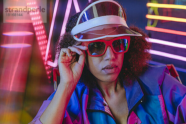 Young woman wearing retro visor and sunglasses in front of neon light