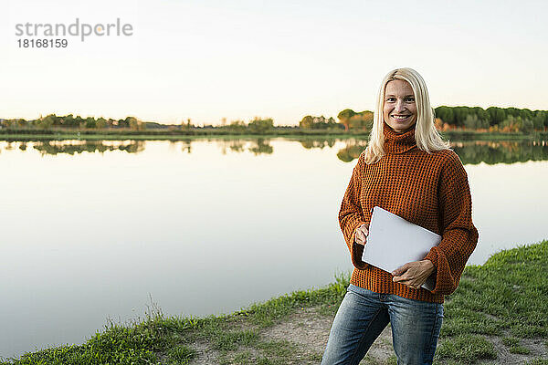 Lächelnder reifer Freiberufler mit Laptop  der in der Nähe des Sees steht
