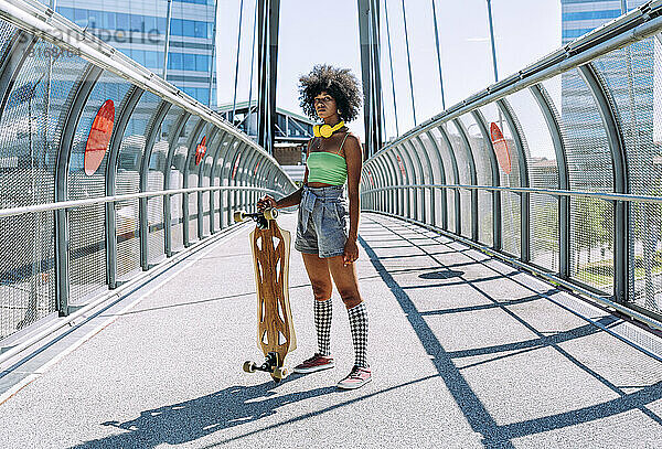 Selbstbewusste junge Frau steht mit Skateboard auf der Brücke