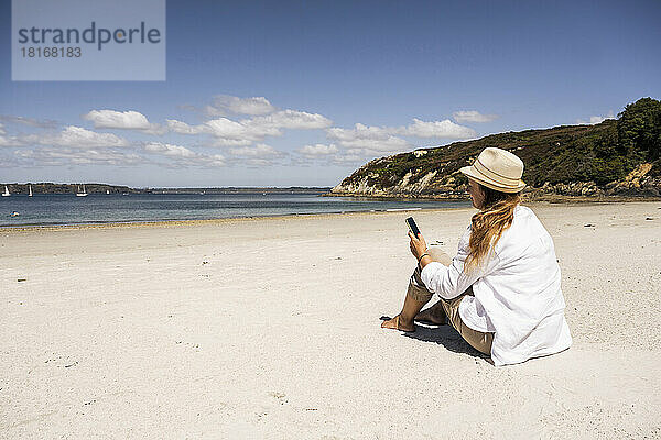Reife Frau benutzt an einem sonnigen Tag ihr Mobiltelefon am Strand