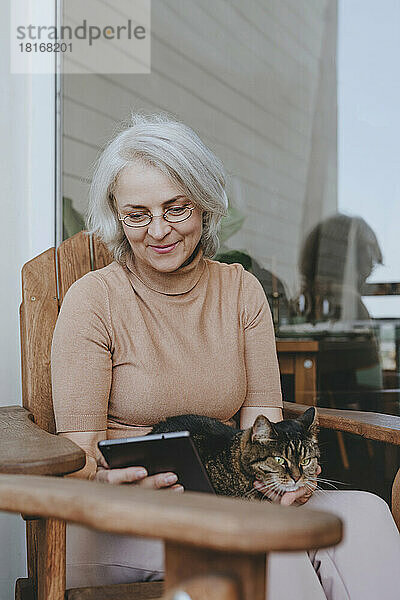 Lächelnde reife Frau benutzt Tablet-PC mit Katze auf dem Schoß auf der heimischen Terrasse