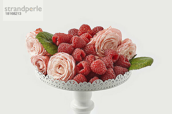 Fresh raspberries with pink roses on pedestal cakestand against white background