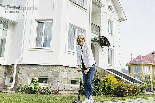 Lächelnde Frau mit Tretroller steht vor dem Haus