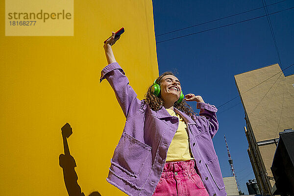 Fröhliche Frau mit erhobener Hand  die an einem sonnigen Tag in der Nähe der Wand Musik hört