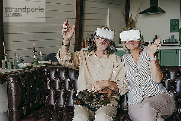 Älteres Paar mit VR-Brille gestikuliert mit Katze auf dem Schoß zu Hause