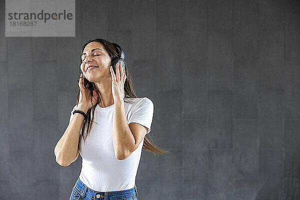 Smiling woman enjoying music through wireless headphones in front of gray wall