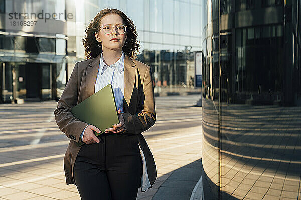 Selbstbewusste Geschäftsfrau läuft mit Laptop am Bürogebäude vorbei