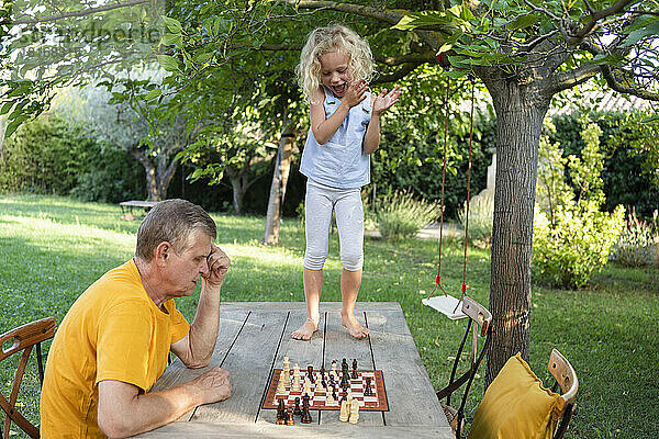 Nettes Mädchen klatscht in die Hände seines Großvaters  der auf dem Tisch im Garten Schach spielt
